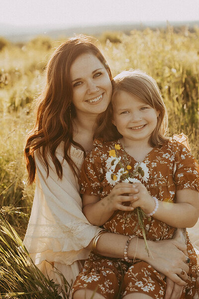 A daughter sitting in her mothers lap holding wildflowers in a beautiful field