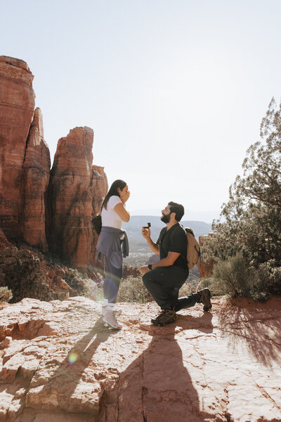Man getting down on one knee to propose to his girlfriend wearing Lululemon workout clothes on top of Cathedral Rock in Sedona, Arizona.