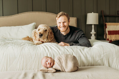 A cozy family moment captured indoors, with a mother and father cradling their newborn baby near a softly lit window. The father gently kisses the mother’s head while she looks down lovingly at their sleeping baby. The natural light enhances the warmth and tenderness of the scene.