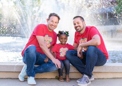 Two gay dads and their little girl smiling in front of a fountain at Creekside Gahanna.