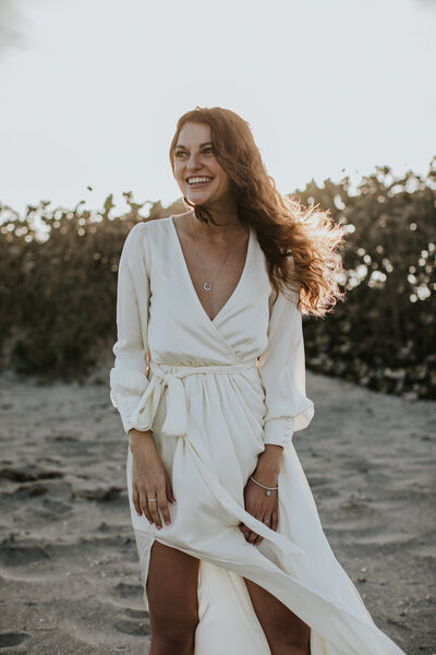 Portrait of a woman on the beach at golden hour with a long white dress blowing in the wind.