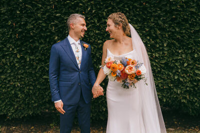 Bride and groom in front of  greenery holding hands and smiling at each other