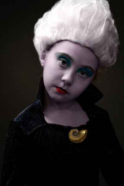 Young girl dressed in Ursula costume with purple makeup on her face, standing in front of black backdrop with somber look.