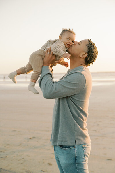 dad holding baby boy up on the beach and giving him a kiss on the cheek