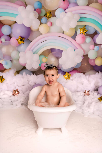 Portrait Photographers CT create magical first birthday bath photos with baby in vintage tub surrounded by pastel rainbow balloons and clouds