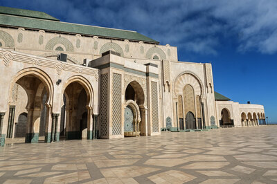 Ornate arches and carved stone details of the Hassan II Mosque in Casablanca, with geometric tilework and a wide open courtyard under a bright blue sky.