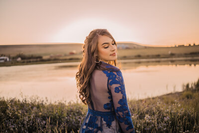 senior girl with long dark hair looking over her shoulder in a blue lace dress next to a pond at sunset