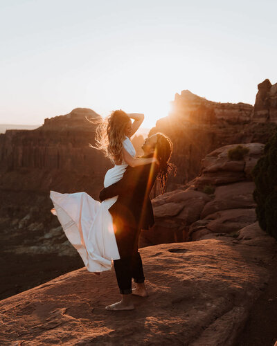 groom holding bride in the air while standing on a rock formation in the desert