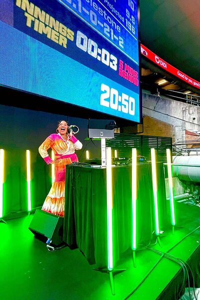 DJ in a vibrant rainbow dress performing beside a glowing DJ booth under a massive live sports scoreboard