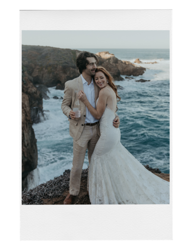 Bride and Groom standing on rock on the coast overlooking the ocean the bride is hugging the groom and leaning against his chest while the groom has his arm around her lower back and is holding a bottle of champagne in the other hand
