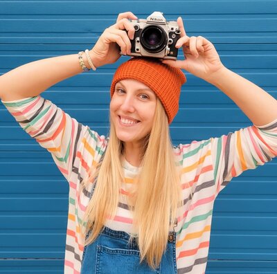 Jessi Cabanin Cadence Photography testimonial photo.  Cadence is light skinned with  long, blonde hair.  She is wearing an orange hat and is holding a camera up on her head.  She is standing in front of a blue  wall.