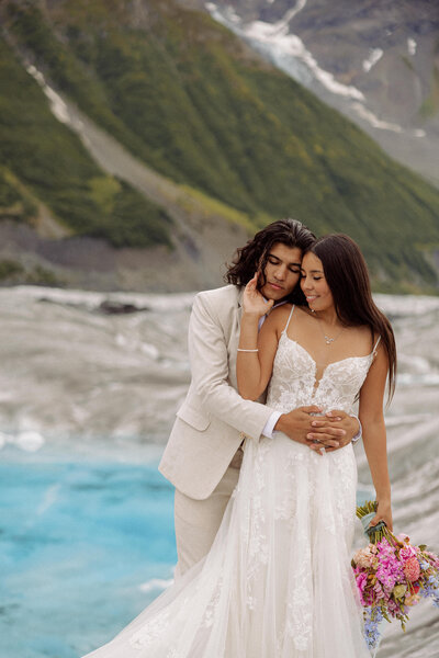 A person hugging their partner from behind as they stand in front of a mountain lake 