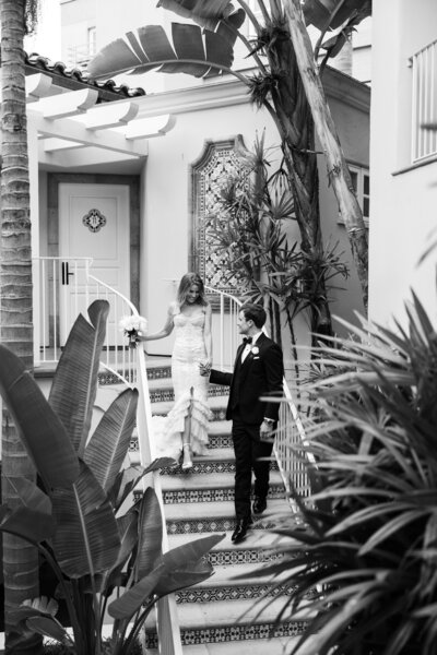Black-and-white photo of bride and groom walking gracefully down a staircase at La Valencia Hotel in La Jolla, San Diego, CA, showcasing their elegant attire against the backdrop of the hotel's iconic Spanish-style architecture and warm, inviting ambiance.