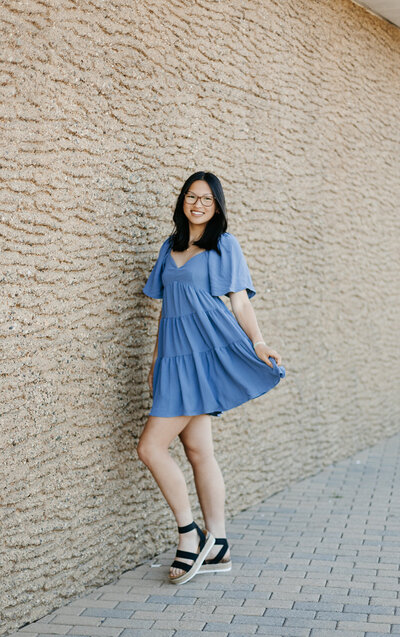 Senior girl in a blue dress and glasses posing outside in front of a stone wall