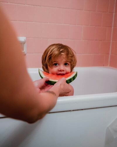 A young child is in a bath, in a pink bathroom. Someone is holding a slice of watermelon in front of the child's mouth.