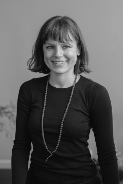 Black and white photo of woman with  short brown hair and bangs wears a black longsleeve and long beaded necklace and smiles at the camera.