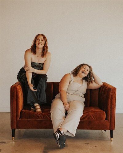 Steph and Andi sit on a burnt-orange couch, laughing.