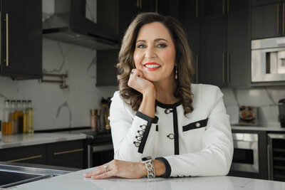 A woman in a white jacket with dark trim smiles while leaning on a kitchen counter with dark cabinets and stainless steel appliances in the background.