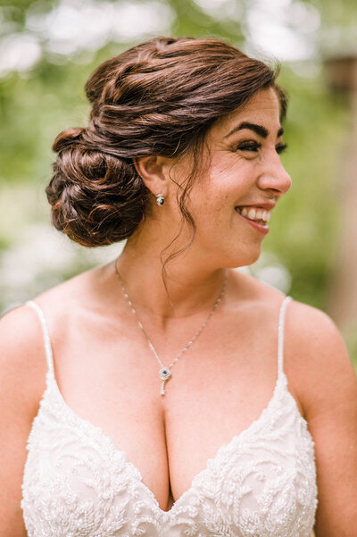 Bride wearing a lace dress with her brunette hair in an updo looks off into the distance while smiling at wedding party at the woods at Algonkian