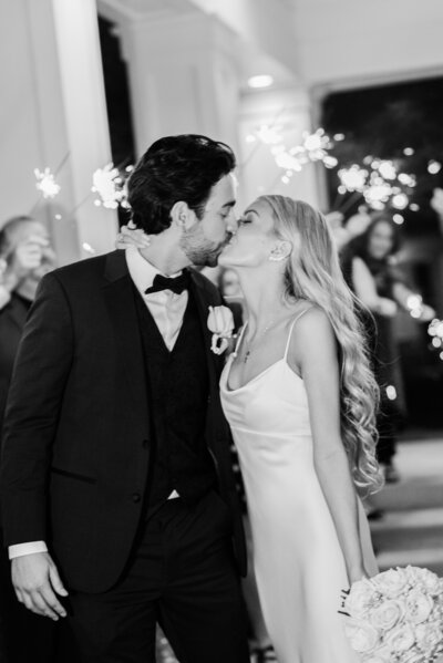 A newlywed couple shares a passionate kiss during their sparkler exit at Sea Pines, Hilton Head Island. The groom in a classic black tuxedo and the bride in a sleek satin gown create a timeless and elegant moment as family and friends celebrate with sparklers.