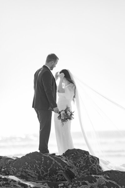 Bride adjusting veil on the beach with groom holding wedding bouquet