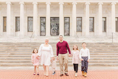 A family of six hold hands and walk together in front of a classical style building