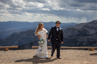 Bride and groom hold hands atop a mountain overlook, with vast rugged peaks and dramatic skies behind them.
