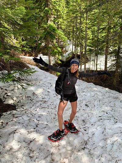 A woman wearing snowshoes walks along a snowy trail surrounded by trees and a clear blue sky.