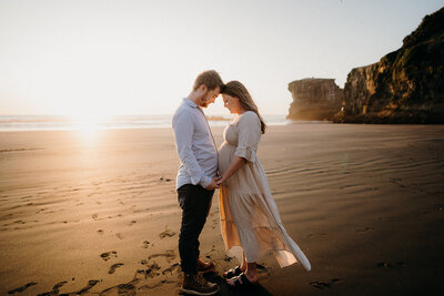 A couple standing with the sunset and the cliffs at Maukatia Bay in Auckland during their maternity photoshoot with Tashina Narelle Photography