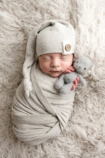 Newborn boy sleeps while holding onto a gray fuzzy tiny stuffed bear, wearing a light gray wrap and gray sleepy cap during his newborn session in Pittsburgh, PA