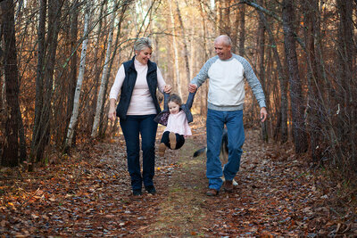 A candid moment of a Grandfather and Grandmother walking down a wooded trail joyfully swinging their granddaughter .