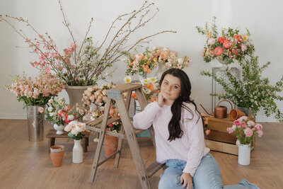Michaela sits on the floor leaned up against a small wooden ladder in front of a floral set up in a photography studio in Bristol, TN