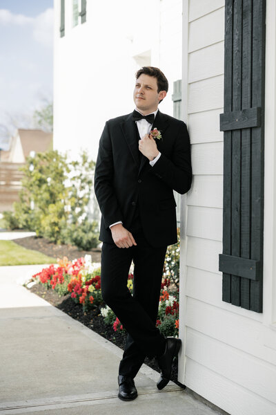Groom in black tuxedo posing outside Maison De Veaux in Houston, Texas with floral boutonniere and spring wedding details in the background