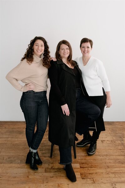 Studio portrait of Canvas Weddings team with owner Corinne in the middle, sitting on stool in front of white background
