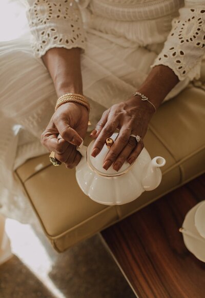 A woman's hand gently rests on a white teapot, capturing the moment before ceremony begins.