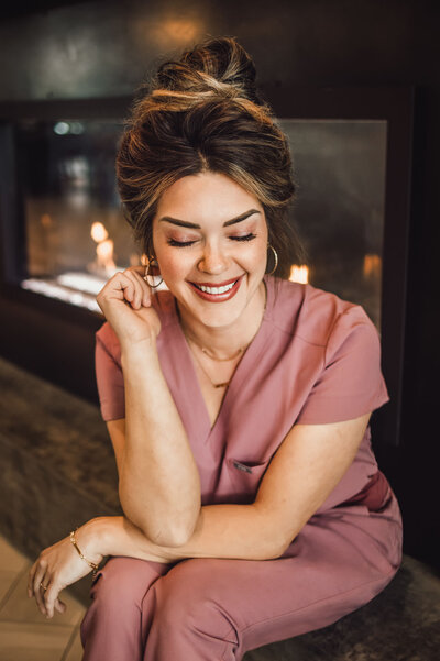 woman sitting in front of fire place wearing pink scrubs while smiling with her hair in a bun