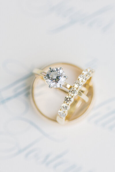 Brilliant cut yellow gold diamond ring with wedding bands sits on top of invitation with blue script font