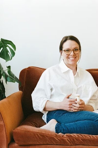 A woman wearing glasses and a white shirt sits on a couch, representing a therapist in Evanston, IL.