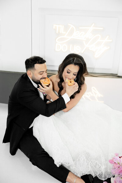 Bride and groom sharing donuts while sitting under neon "'Til Death Do Us Party" sign at Maison De Veaux in Houston, Texas during playful wedding reception moment