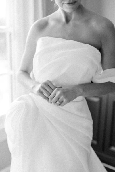 Elegant black-and-white portrait of a bride adjusting her gown, captured at Winbrooke in Brookshires, Massachusetts. The image highlights the bride's delicate hand placement on her dress, showcasing the intimate, thoughtful moments of wedding preparation. Felisha, a New England-based destination wedding photographer, captures timeless, authentic wedding moments in Massachusetts, New England, and worldwide locations.