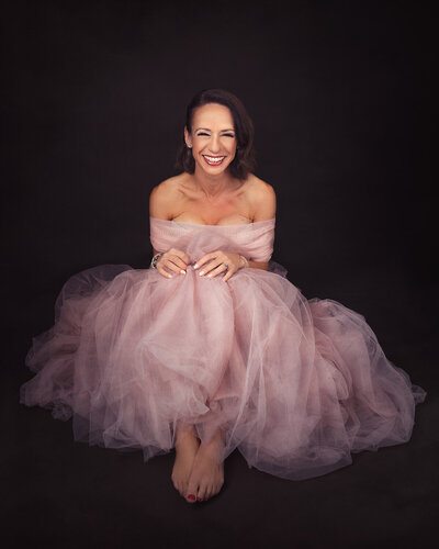 Woman in a pink tulle dress sitting on the floor and smiling to the camera. Taken in the studio on a black background by Norwich Photographer, Claire Howes.