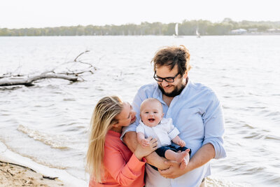 A family stands by the water's edge, with the father holding their smiling baby while the mother leans in to kiss the baby.