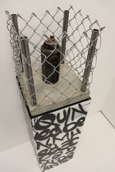 A dark ceramic spray paint can is decorated with a carving of young children and sits atop a graffiti painted pedastal, but is caged in by a wire fence