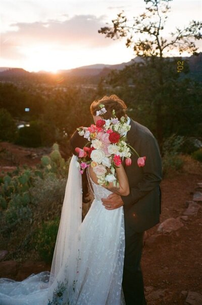 A couple shares a kiss while hidden behind a large bouquet of flowers during their wedding in Sedona, Arizona. The vibrant sunset colors the sky as they embrace, with desert flora visible around them.