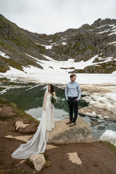 Bride and groom share a quiet moment by a glacier and alpine water, surrounded by rugged mountain peaks and late-spring snow.