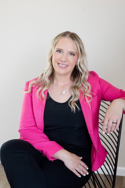 Becca sitting in a chair wearing a bright pink blazer, smiling.
