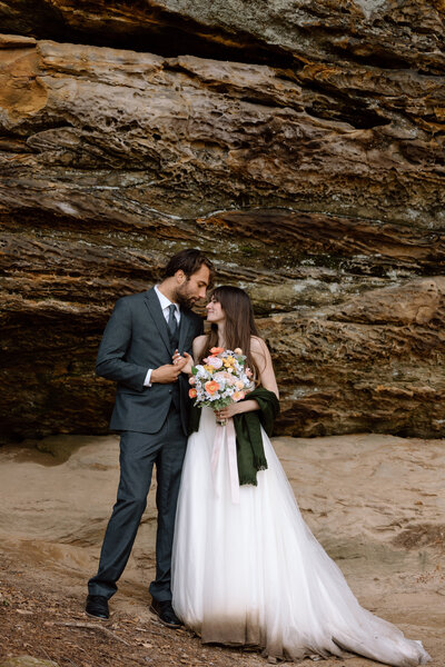 Couple standing side by side and looking at each other, interesting rocks are behind them.