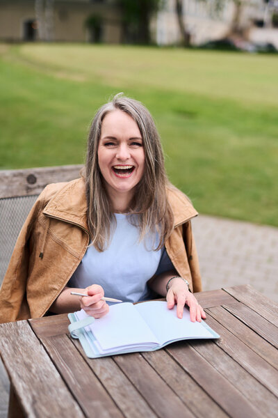 Renata Jaroscakova from Real Joy Photography sitting at the table in a garden, writing in her notebook and laughing