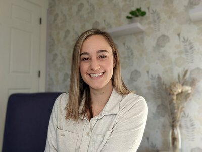 Lexi smiles happily at the camera, with shoulder-length blonde hair resting on her shoulders, wearing a tan, fleece button-down top. Behind her, the wall is covered in a plant-patterned wallpaper, and a small houseplant is visible on a shelf above her head.