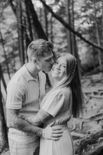 Couple embrace with man kissing woman's cheek in a forest.